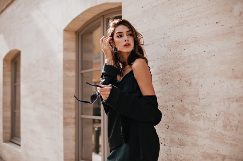 Home cute young girl with dark wavy hairstyle bright makeup silk dress black jacket holding sunglasses hands looking away against beige building wall 1.jpg