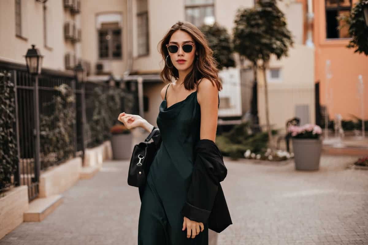 Home fashionable pale brunette long green dress black jacket sunglasses standing street during daytime against wall light city building 1.jpg
